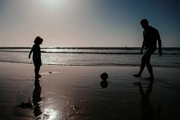 父と子供は屋外で楽しんでいます 父と息子は夏の家族の休日にビーチでサッカーやサッカーをし 日没のシルエット 幼少期と育児の概念 — ストック写真