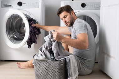 Çamaşır makinesinin yanında elbiseli bir adam. Yakışıklı adam çamaşır makinesinin önünde oturur. Çamaşır makinesini kirli çamaşırlarla dolduruyor. Adam elbiseleri temizliyor. Yalnız bir adam için ev işi. Ev çamaşırları