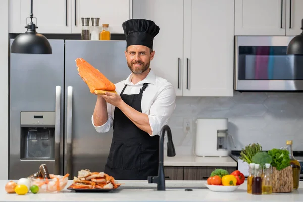 Portrait of cook man preparing fresh salmon at kitchen. Handsome man in cook apron and chef hat preparing raw fish salmon. Chef cooking seafood in kitchen. Millennial man hold raw fish salmon