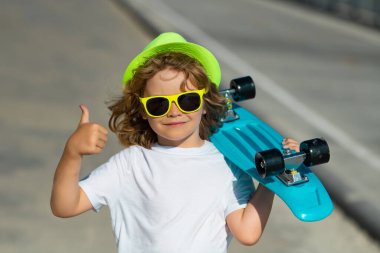 Çocuk sokakta uzun tahta tutuyor. Pennyboard 'u açık havada olan çocuk. Bozuk paralı neşeli küçük bir çocuğun açık hava portresi. Heyecanlı çocuk başparmağı havada, sokak portresi yakın.