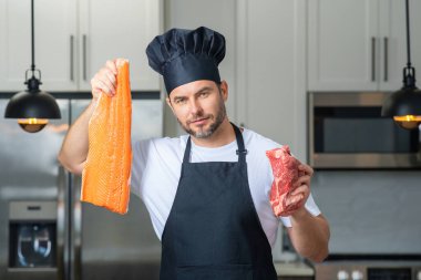 Aşçı adam balık ve et, somon ve sığır eti tutuyor. Aşçı üniformalı, çiğ et ve balık fileto balıklı bir aşçı. Aşçı mutfakta çiğ et ve balık filetosu pişiriyor.