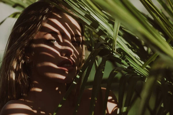 Closeup portrait of beautiful woman with shadows of palm leaf on her face. Beautiful young woman with nude makeup looking trought palm leaves