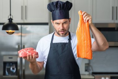 Aşçı adam balık ve et, somon ve sığır eti tutuyor. Aşçı üniformalı, çiğ et ve balık fileto balıklı bir aşçı. Aşçı mutfakta çiğ et ve balık filetosu pişiriyor.