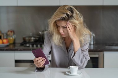 Sabah mutfakta bunalımlı bir kadın. Mutfakta kahveli ve telefonlu üzgün bir kız. Güzel bir kadının sabahı. Akıllı telefon kullanan üzgün bir kadın.