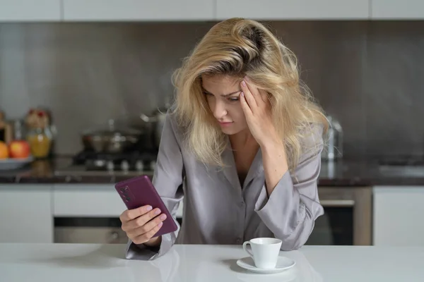 Depressed woman in the kitchen in the morning. Upset girl with phone with coffee in kitchen. Morning of pretty woman. Sad lady using smartphone