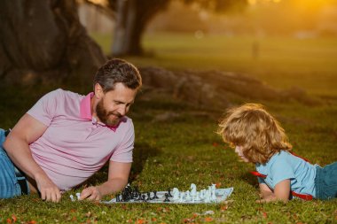 Baba oğul çim parkında çimlerde satranç oynuyorlar. Genç bir çocuk satrançta bir adamı yeniyor. Babalar Günü, aileyi sevmek, ebeveynlik, çocukluk kavramı.
