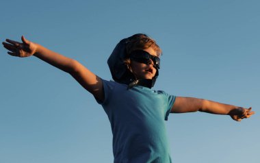 Pilot gözlüklü ve kasklı mutlu çocuk mavi gökyüzü arka planına karşı oynuyor. Güneşli bir yaz gününde gökyüzünde yürüyen sevimli bir çocuk. Çocuklar seyahat hayali kurar.