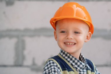 Elinde çekiç tutan kasklı bir çocuk. Mutlu gülümseyen küçük çocuk inşaat alanında oyuncak aletlerine yardım ediyor. İnşaat aletleri olan çocuklar. İnşaat işçisi