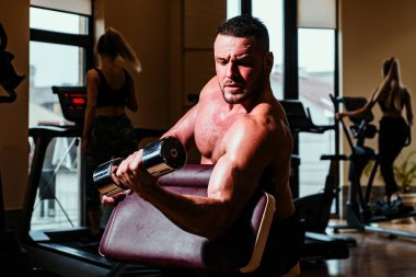 Spor salonunda dambılla egzersiz yapan genç sporcunun portresi. Dambıllı adam eğitimi. Kaslı adam dambıllarla egzersiz yapıyor. Dambılı kaslar