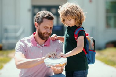 Çocuklar için okul yemeği. Baba oğlunu destekliyor ve motive ediyor. Okul çocuğu ve ebeveyni tişörtlerinde beslenme çantası tutuyorlar.