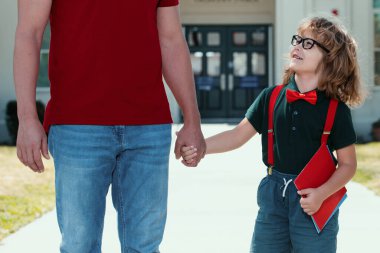 Öğretmenlerin elini tutan mutlu inek öğrenci portresi. Amerikalı baba ve oğul okul parkında yürüyorlar.