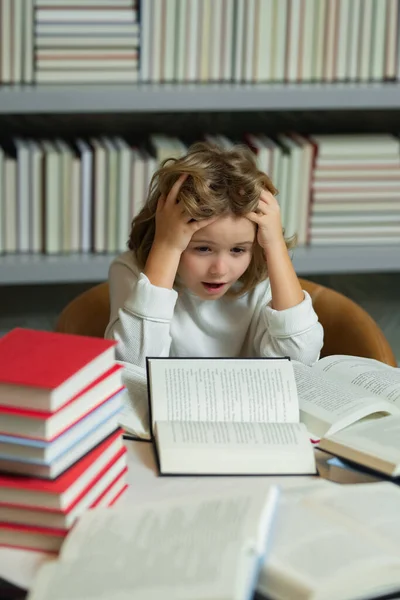 stock image Intellectual child, clever pupil. Child boy doing homework on desk in school library. Schoolboy studying homework literature near pile of books. Kid reads a book with shelves stacks of books