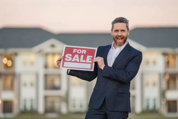 Handsome real estate agent holding banner house for sale. House with land and insurance. Man real estate agent in business suit presenting the house for sale. Land agent trying to sell family house