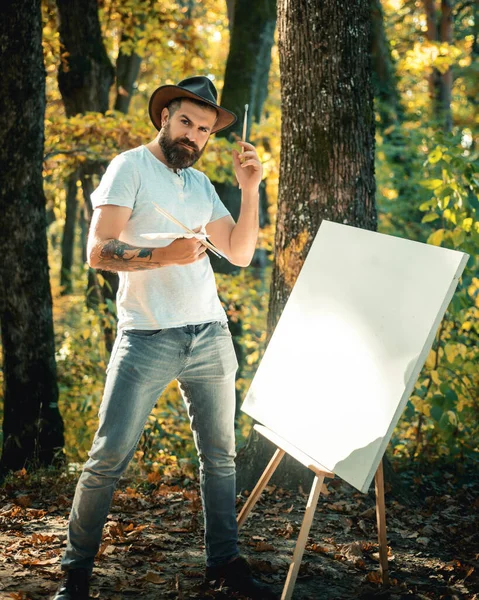 Hipster artist in a hat creating art in the woods. Art concept. Painting in nature. Start new picture. Painter with easel and canvas