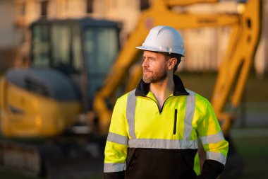 İnşaat alanında miğferli bir işçi. Kazıcı buldozer erkek işçi. Arkasında kazıcı olan inşaat işçisi. İnşaat işçisi traktörlü ya da inşaatta çalışıyor.