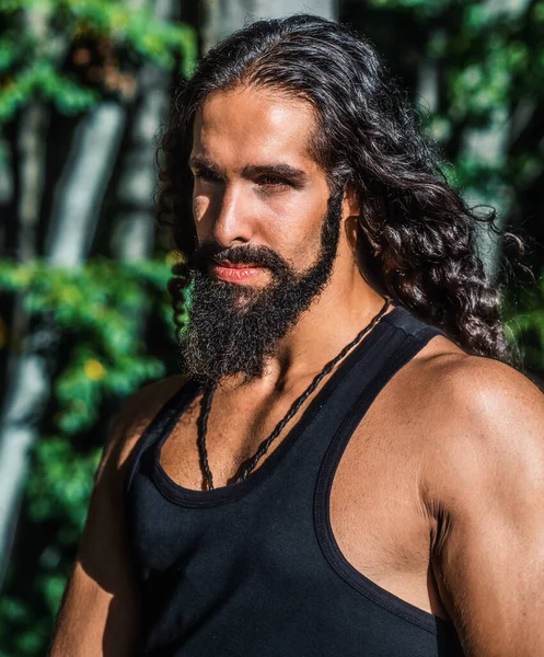 Handsome bearded man with long dark hair. Sexy bearded model man. Man fashion and style concept. Portrait of handsome young strong man with long dark hair looking away while standing at forest