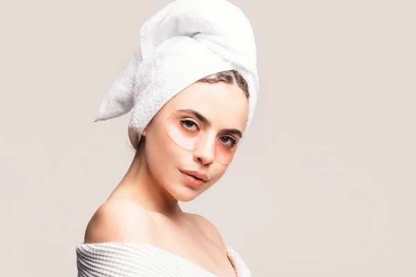一个头戴毛巾 身穿白袍的年轻漂亮的姑娘 用眼皮底下的补丁来照顾皮肤 使用抗疲劳面罩 女孩减少眼袋 — 图库照片