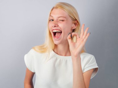 Göz kırpan güzel bir genç kadının portresini yakından çek. Göz kırpan sevimli, komik bir bayan. Beyaz arka planda izole edilmiş bir kadın parmaklarıyla işaret gösteriyor. Komik genç kız güzel jest yapıyor.