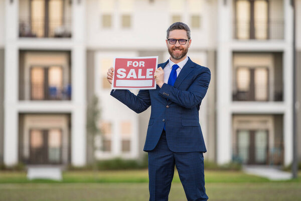 Успешный риэлтор в костюме с вывеской на продажу. Риэлтор перед новым домом. Продавец продает дом, новую квартиру в современном доме