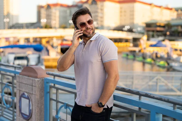 Portrait of handsome man chatting on phone in city. Confident man talking on phone in modern big american city. Stylish lambersexual model. Sexy man dressed in polo. Bisiness phone conversation