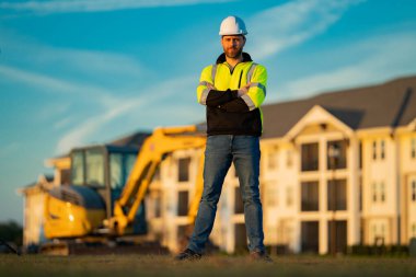 Worker in helmet on site construction. Excavator bulldozer male worker. Construction driver worker with excavator on the background. Construction worker with tractor or construction at building