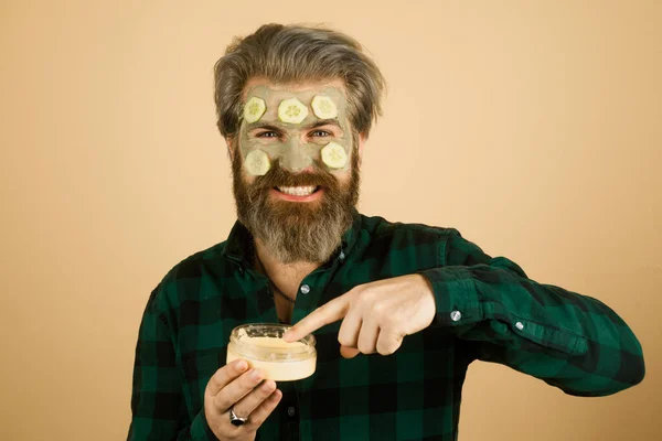 Facial mask. Man getting facial care with cucumber. Close up portrait of a laughing bearded man applying face cream mask and looking in camera, isolated over yellow background