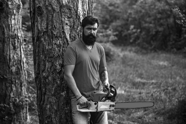 森のチェーンソー付きの木材ジャック労働者 森の近くに斧を持つハンサムな若い男 スタイリッシュな若い男は腰椎のようにポーズ 髭を生やしたハンサムな若い男が木を持っている — ストック写真