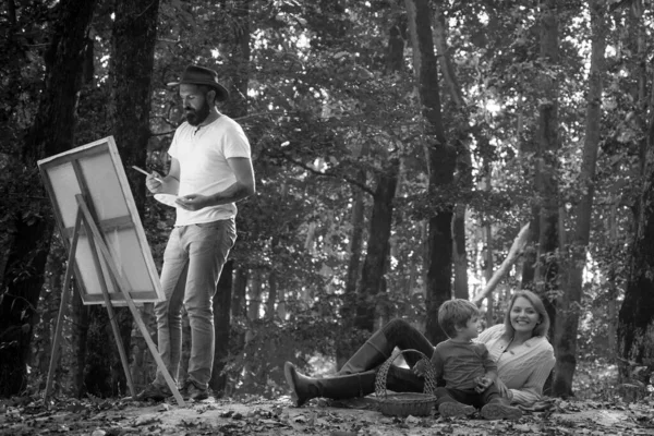 ハンサムな髭の男は 絵具とへらでパレットを使用して公園で彼の若い家族の写真を描きます 密接な関係者の写真とイーゼルとキャンバスを描く — ストック写真