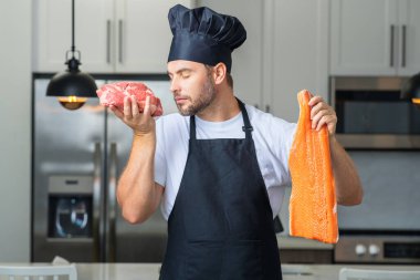 Aşçı, mutfaktaki evde taze doğal yemek hazırlıyor. Yakışıklı neşeli adam çiğ balık, et, somon ve biftek hazırlıyor. Sağlıklı yemekler, yemek pişirme konsepti. Somon balığı fileto bifteğe karşı.