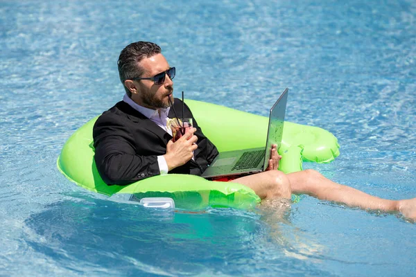 Summer business dreams. Business man in suit floating with cocktail and laptop in swimming pool. Summer business vacation. Funny crazy businessman rest in formal wear in swim pool. Hot summer business