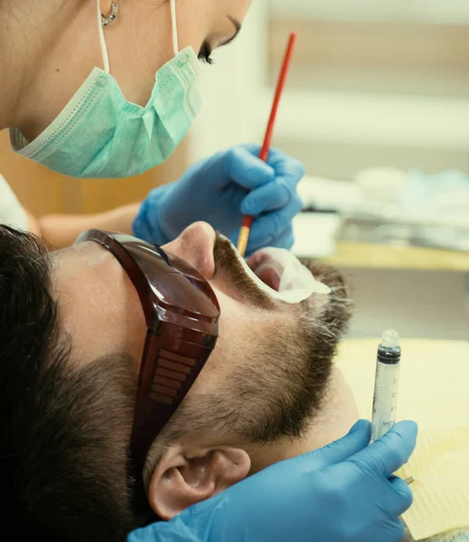 牙医在光线模糊背景上检查年轻人 在现代牙科诊所用镜子检查病人牙齿的女牙医 胡子男子在牙医那里检查牙齿 — 图库照片
