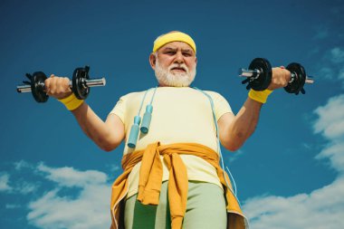 Kıdemli sporcu mavi gökyüzü arka planında dambıl kaldırarak egzersiz yapıyor. İzole edilmiş, kopyalama alanı. Spor salonundaki son sınıf öğrencisi ağırlık çalışıyor. Kıdemli adam mavi gökyüzünde egzersiz yapıyor.