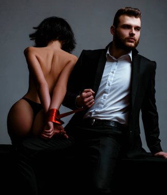 Seksi kız çıplak kalça. Tutkulu aşıklar. Seks hakimiyeti. Seks teslimiyeti. Maço bakımlı hipster ve çıplak seksi kadın elleri kırmızı kurdele bağlı. Cinsel ilişkiler. Seks oyunu. Tease ve baştan.