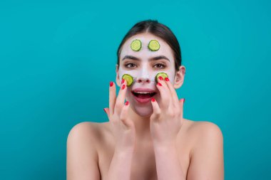 Close up beauty portrait of a beautiful woman applying face cream and facial mask of cucumber. Mask for skin lifting and anti-aging effect, studio background. Anti aging cream. Facial treatment