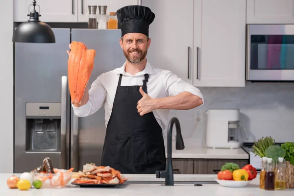 Portrait of cook man preparing fresh salmon at kitchen. Handsome man in cook apron and chef hat preparing raw fish salmon. Chef cooking seafood in kitchen. Millennial man hold raw fish salmon