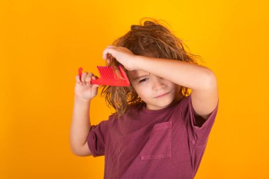 Sarı stüdyo arka planında izole edilmiş saçlarını tarayan küçük çocuk portresi. Taraklı şirin bir çocuk. Sarışın çocuk asi saçlarını tarıyor. Uzun saçlı çocuk.