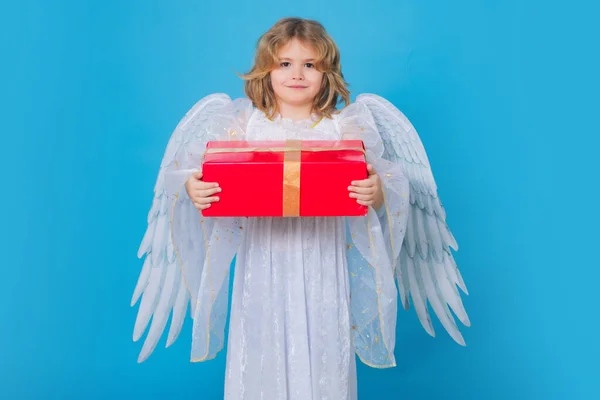 Little angel with gift box present. Valentines day. Blonde cute child with angel wings on a blue isolated studio background. Happy angel child