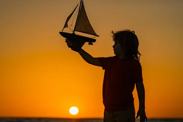 日没の海でおもちゃのセーリングボートで遊んでいる子供のシルエット 小さなブロンドの少年は夏休みの間 ビーチで海の波におもちゃのボートを置きました セーリング船での旅行ツアー 子供の夢のコンセプト — ストック写真