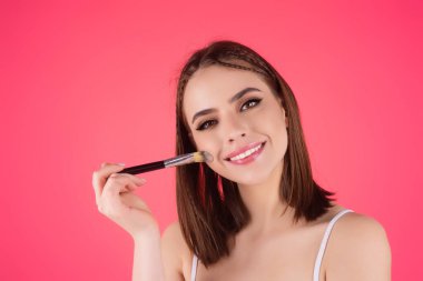 Beautiful young woman applying makeup on studio background. Pretty girl holding makeup brushes and make up on face with cosmetics. Facial beauty. Perfect skin, natural make up