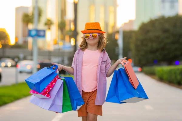 夏のファッションの服でかわいい男の子は買い物に行きます 手にショッピングパッケージと幸せな子供 買い物袋の屋外で買い物をする子供 買い物袋を持って通りを歩くかわいい子供 — ストック写真