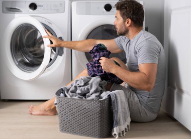 Çamaşır makinesinin yanında kirli giysileri olan seksi bir adamın portresi. Yakışıklı adam çamaşır makinesinin önünde oturur. Kirli çamaşırlar. Adam elbiseleri temizliyor. Yalnız bir adam için ev işi. Ev çamaşırları