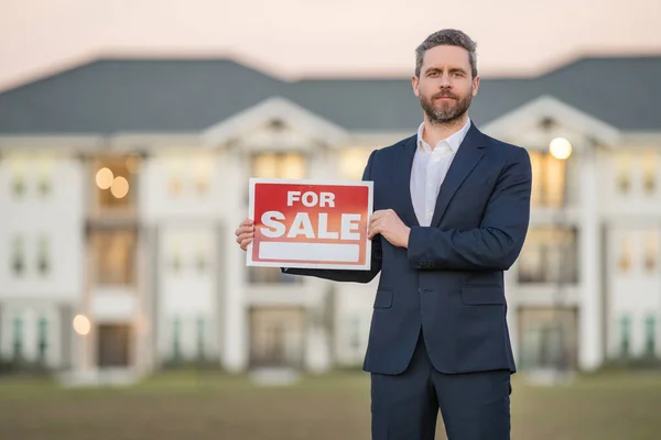 Successful real estate agent in a suit holding for sale sign near new apartment. Real estate agent with home loan contract, selling home. Realtor or real estate agent shows board for sale