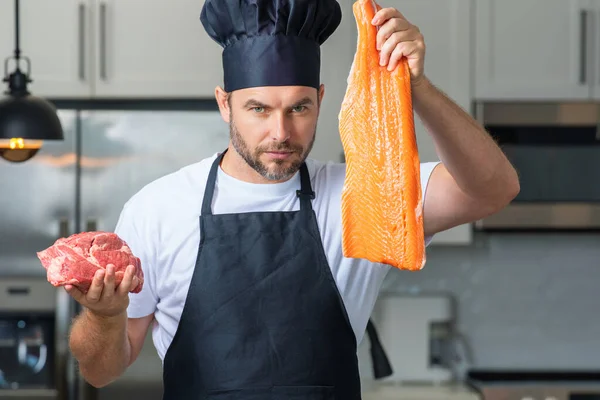 Professional kitchen. Chef with raw meat beef and fish salmon fillet. Man in uniform on kitchen. Handsome male chef in uniform with chef hat cooking raw meat beef and fish salmon fillet in the kitchen