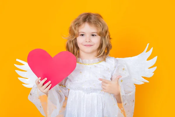 子供の天使紙の赤い心 ポイントジェスチャーを指している 子供の天使 黄色のスタジオの背景に隔離された天使の翼とかわいい子供の肖像画 小さな天使 バレンタインデー 天使のような子供たち — ストック写真