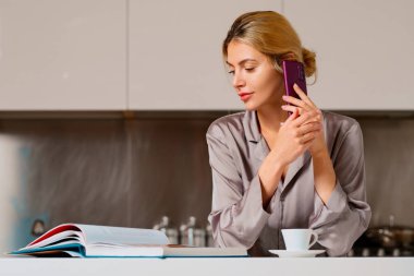 Genç bir kadın cep telefonuyla sohbet ediyor, kitap ya da dergi okuyor ve mutfakta kahve içiyor. Sabahları ev hanımı modundayım.