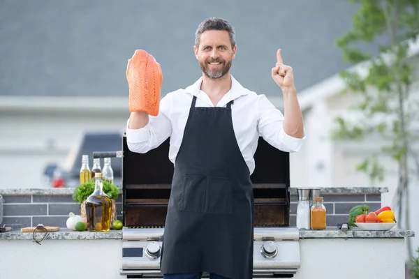 厨师在花园里烤围裙和烤鲑鱼鱼 一个英俊的男人在准备烤鲑鱼片 烤肉和烤肉后院里煮鱼 — 图库照片