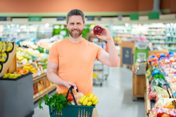 Man with shopping basket full of vegetables and fruits. Middle aged millennial man in a food store. Supermarket shopping and grocery shop concept. Man man 40s with shopping basket