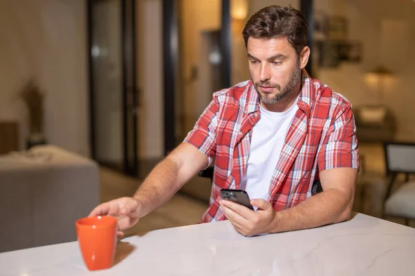 Portrait of man looking at smart phone at living room. Man is talking hold cup of coffe chatting on the smartphone. Thoughtful man using mobile phone at home. Middle-aged man using phone