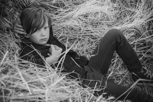 Kid boy holding gold leaf and lies on the hay. Fair-haired boy lies on hay background and eats an apple. Bye summer - hi autumn. Kid on an autumn holiday in the farm