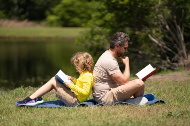 Ot üzerine kitap okuyan sevimli bir aile. Oğlan ve baba parkta piknik yapıyorlar. Mutlu oğul ve baba kitap okuyor. Mutluluk ailesi. Babam parkta çimlerin üzerinde oturan bir çocuğa kitap okuyor.